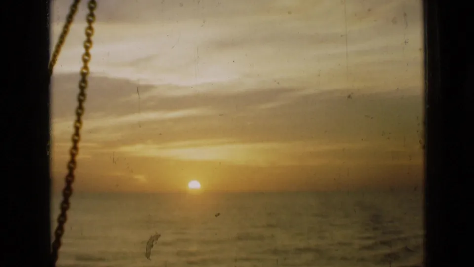 A warm sunset over the sea appears through a dark window opening, with a hanging chain and visible 8mm dust and scratches.