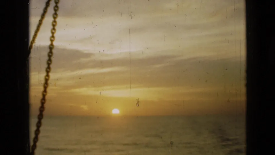 A steady 8mm frame of sunset over water, with warm orange sky, a dark window divider, and a chain silhouetted on the left.