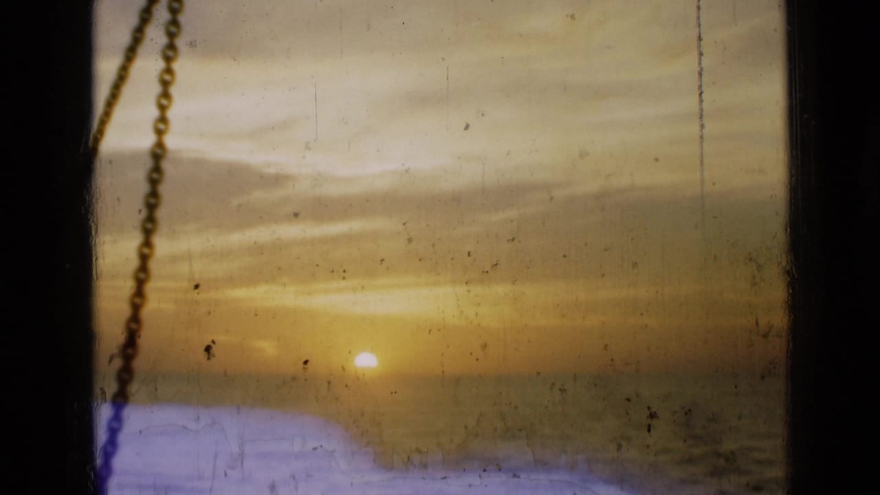 1946 home-movie frame of a low sun over water, viewed through a window or porthole-like opening with a chain at the left.