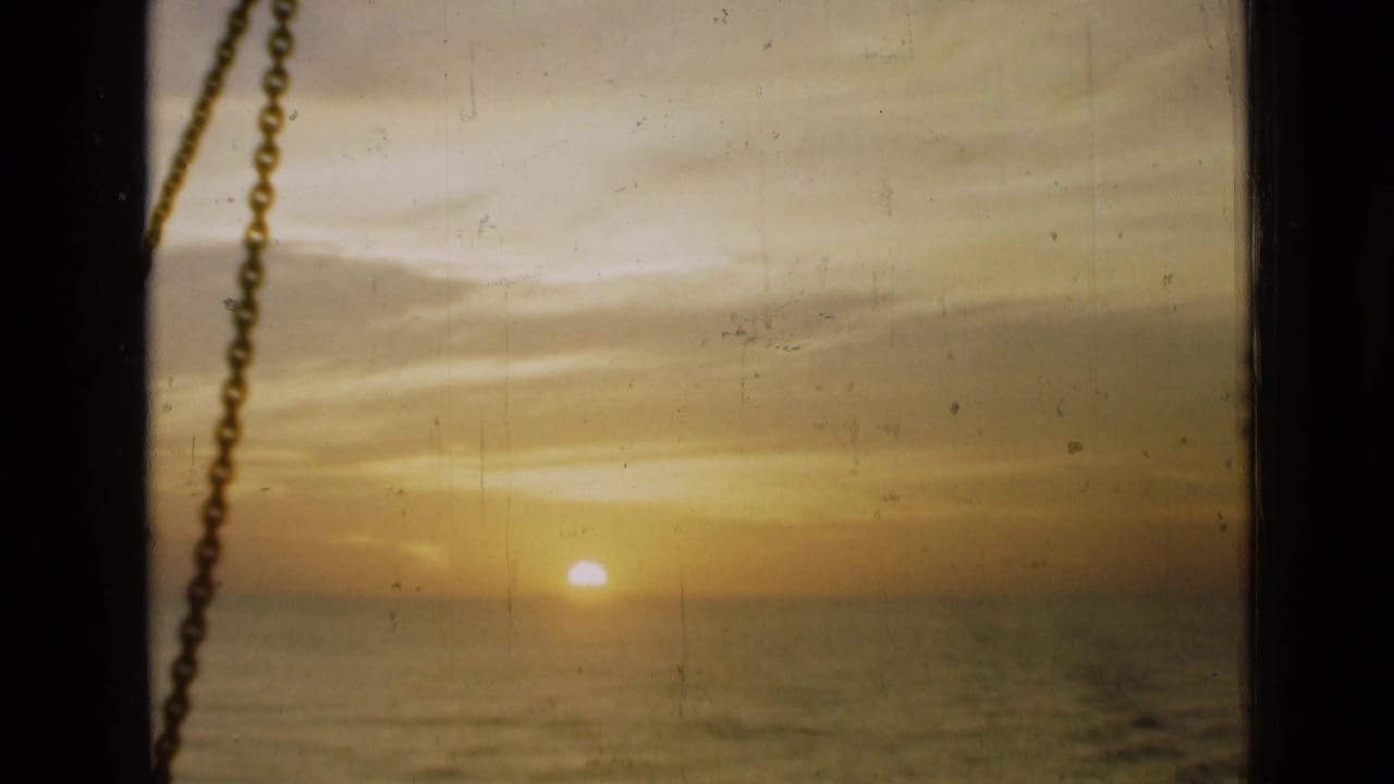 Warm 1946 8mm view of the sun near the water horizon, with dark window edges and a chain visible in the foreground.