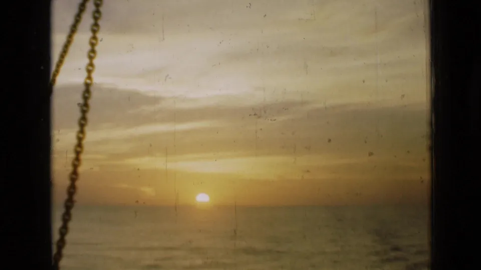 Warm 1946 8mm view of the sun near the water horizon, with dark window edges and a chain visible in the foreground.