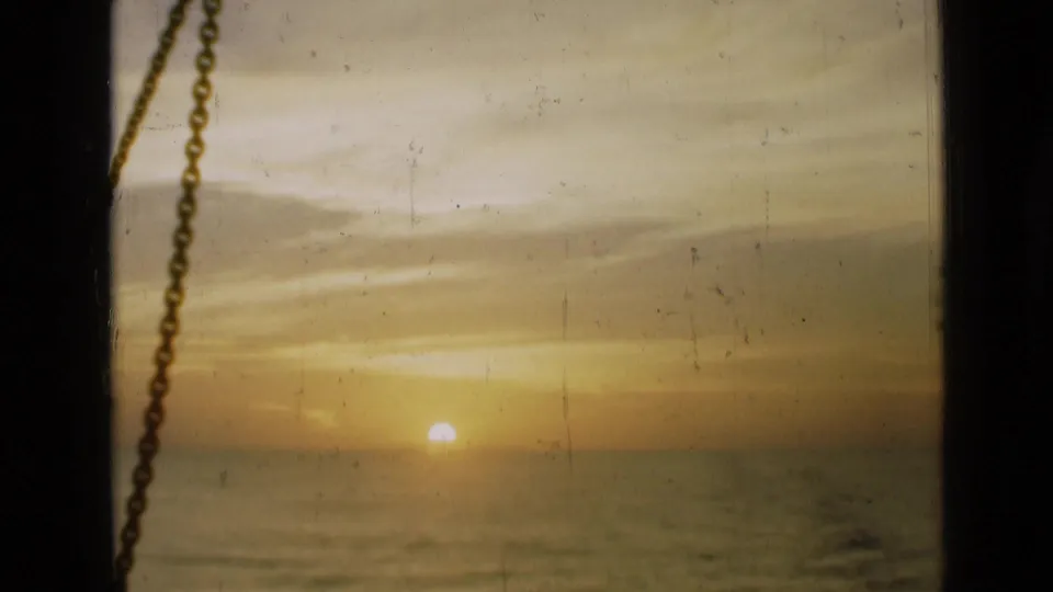 Soft 1940s home-movie frame showing a hazy sunset over water, partially framed by a hanging chain and dark window edge.
