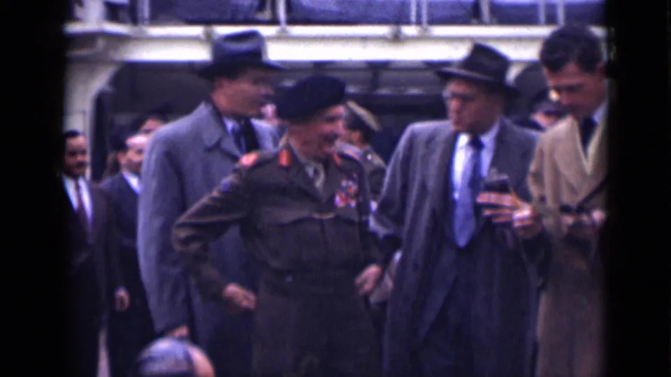 1946 London home-movie frame showing suited civilians and a beret-wearing military officer with medals walking together on a dock or ship deck.