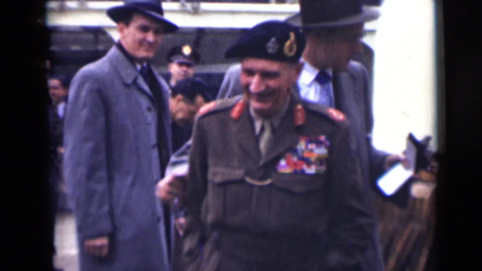 The officer smiles while passing close to the handheld camera, with suited men and uniformed personnel visible behind him in London.