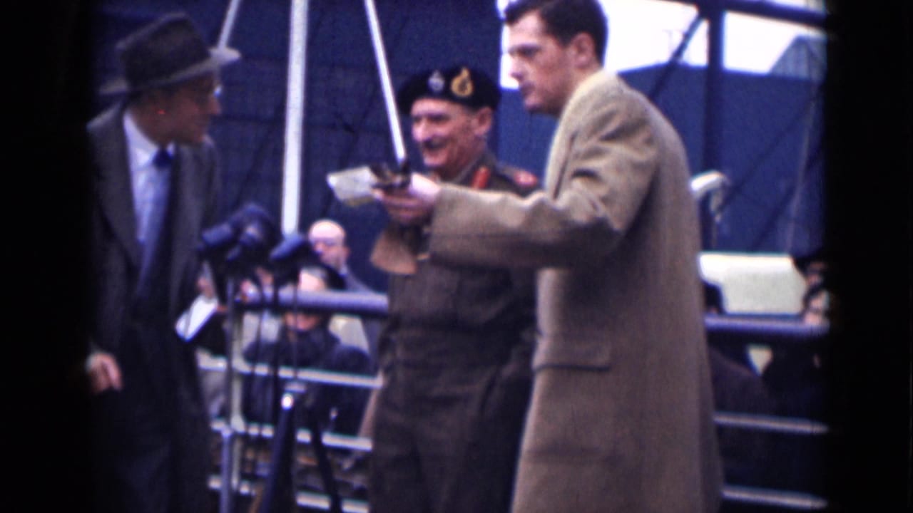 Military officer and suited men stand on an outdoor platform near microphones and spectators.