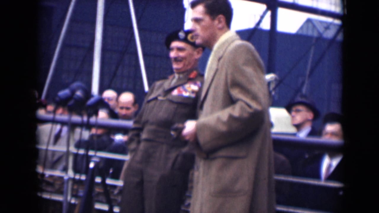 A decorated military officer smiles on a 1946 London platform as a man in a tan overcoat stands beside him, with microphones and spectators visible behind.