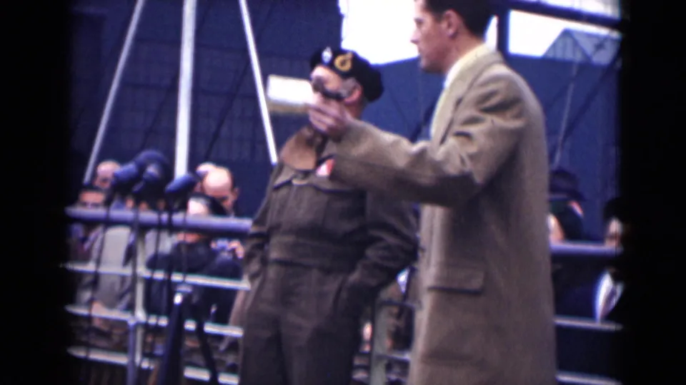 A man in a tan overcoat gestures with a small object on the London platform, while the decorated officer stands close to the microphones.
