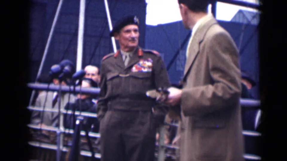 The decorated officer turns toward the man in the tan overcoat on the 1946 London platform, with microphones and seated onlookers behind them.