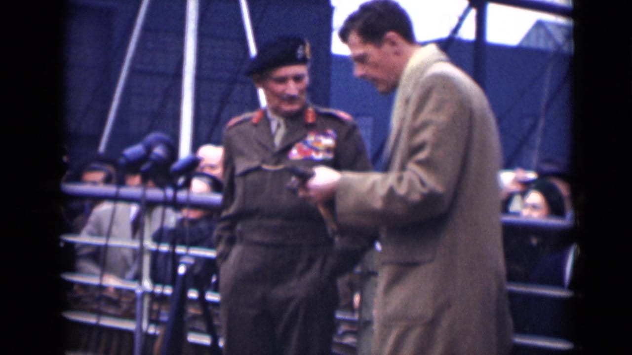Decorated uniformed veteran stands beside a younger man in a beige overcoat at a microphone-lined platform.