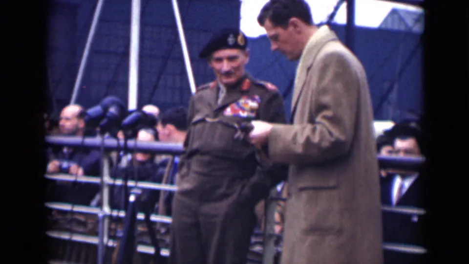 The London 1946 home movie shows the decorated veteran with a cane standing near the overcoated presenter, with onlookers behind a railing.