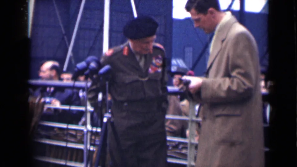 At the end of the London 1946 ceremony clip, the decorated veteran lowers his gaze beside the presenter holding paper near the microphone stands.