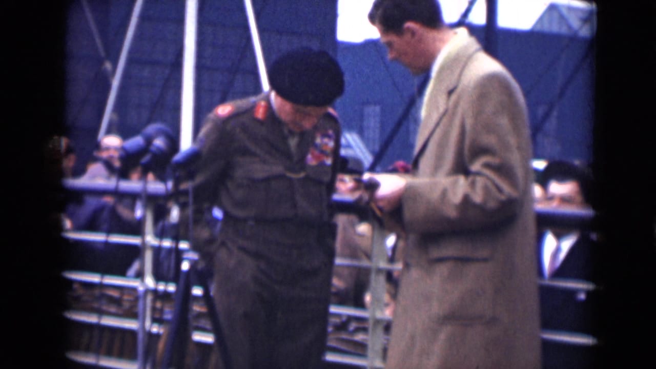 Decorated military officer in a beret stands beside a man in an overcoat near press microphones and railings.