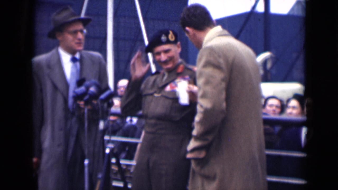 In 1946 London, the decorated officer raises a hand beside clustered microphones as suited men and onlookers stand nearby.