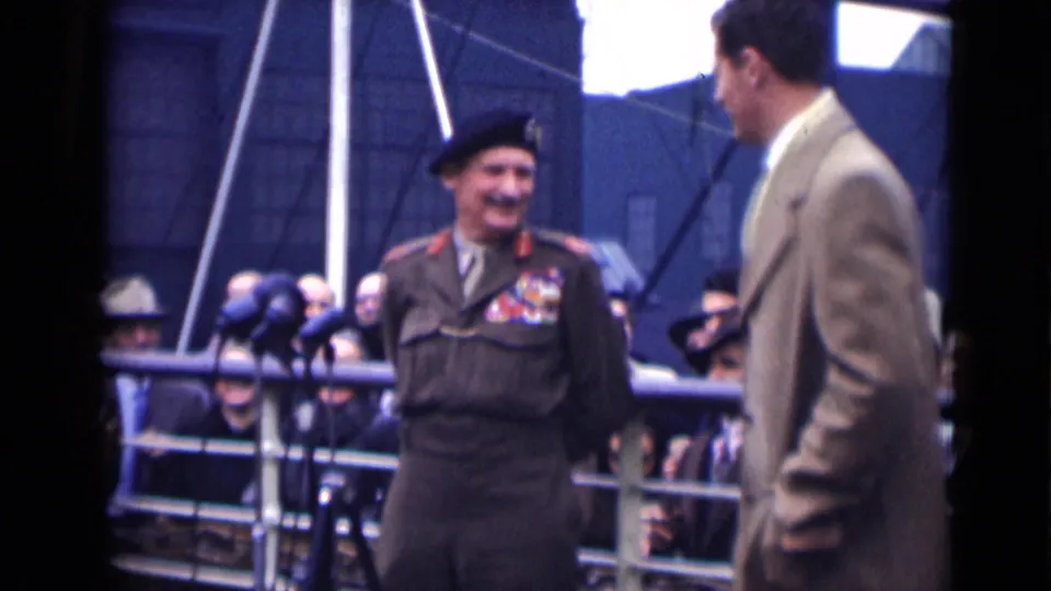 In 1946 London, the officer smiles in uniform beside press microphones while an overcoated man turns toward him.
