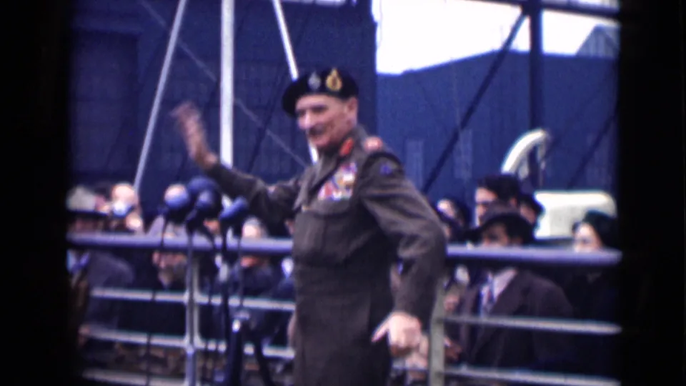In 1946 London, the decorated officer waves broadly near press microphones, with civilians watching from behind metal railings.