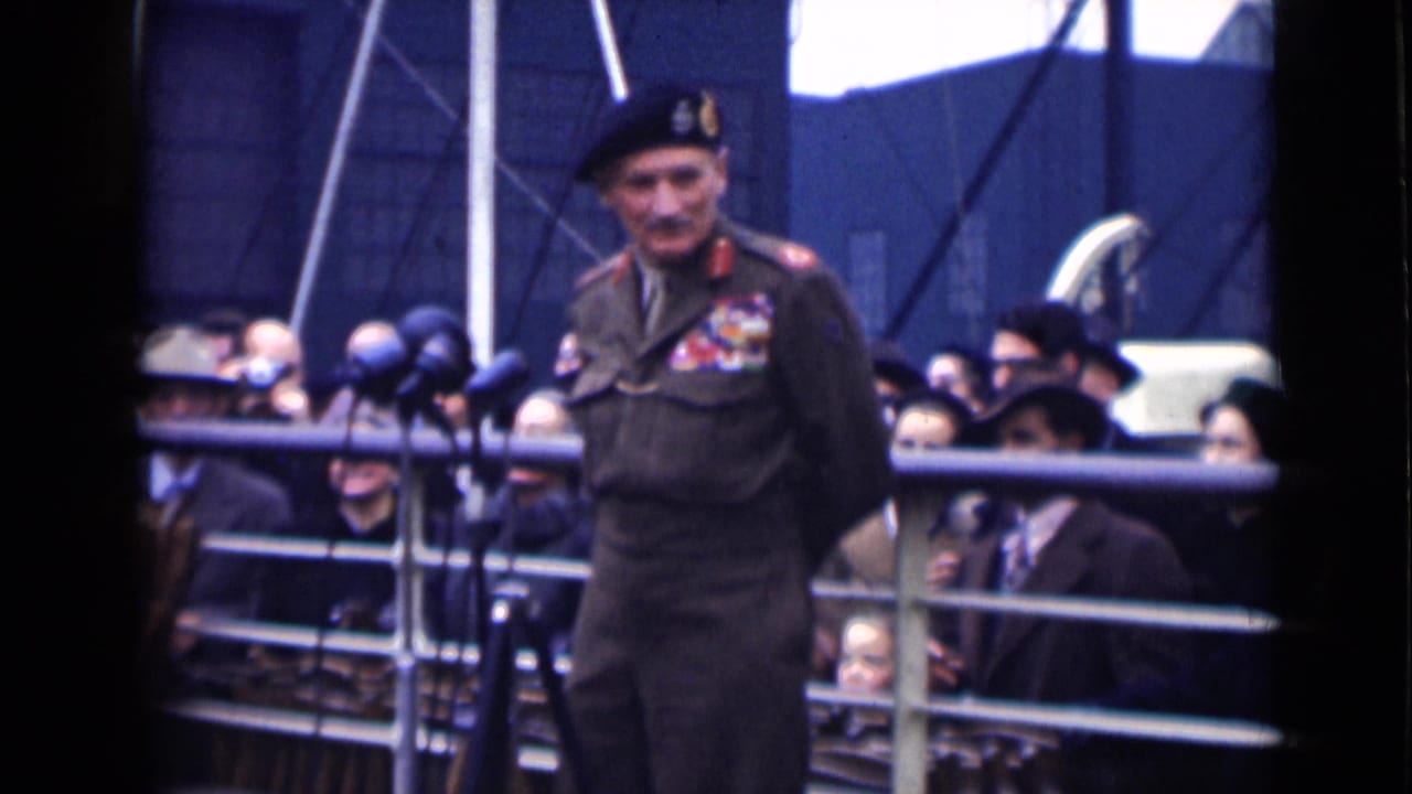 Decorated military officer in beret stands before microphones with a crowd behind railings.