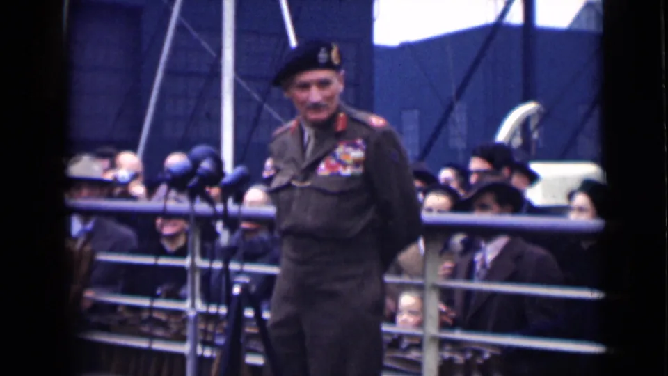 In London, 1946, a decorated military officer in a beret stands at microphones as spectators watch from behind metal railings.