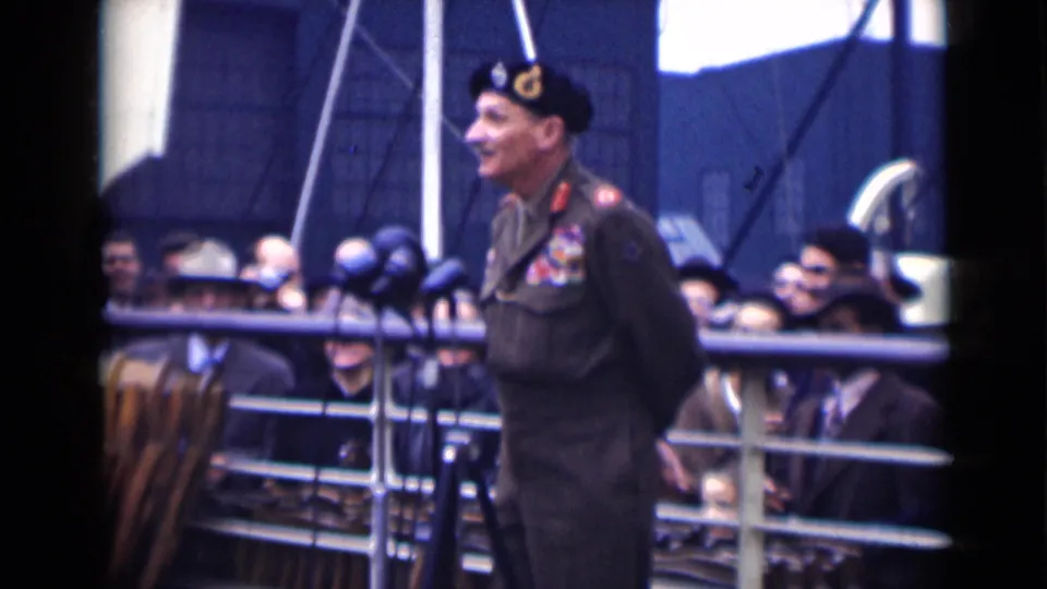 Side view of the decorated officer speaking at a London public event in 1946, with microphones and onlookers visible behind him.