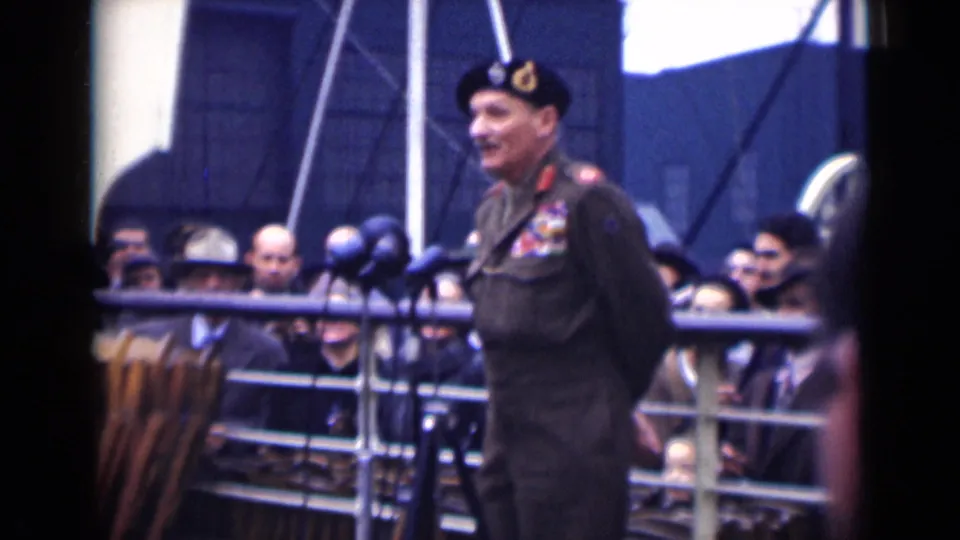The officer remains in profile at the microphone position, with London spectators and railings forming the public-event background.