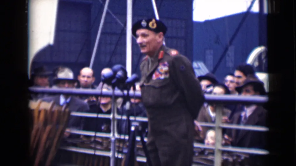 In London 1946 home-movie footage, a decorated uniformed officer stands on a ship deck near microphones as civilians watch behind railings.