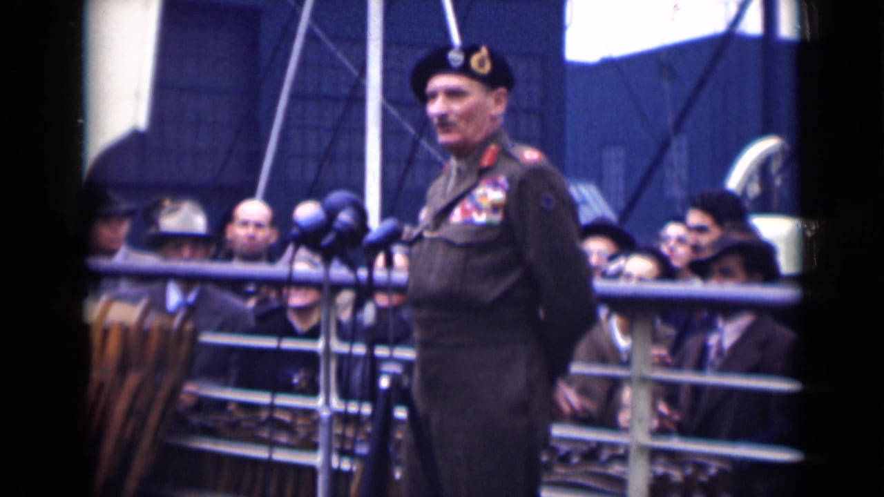 A decorated officer in uniform appears beside several microphones on a deck, with London 1946 spectators visible behind him.