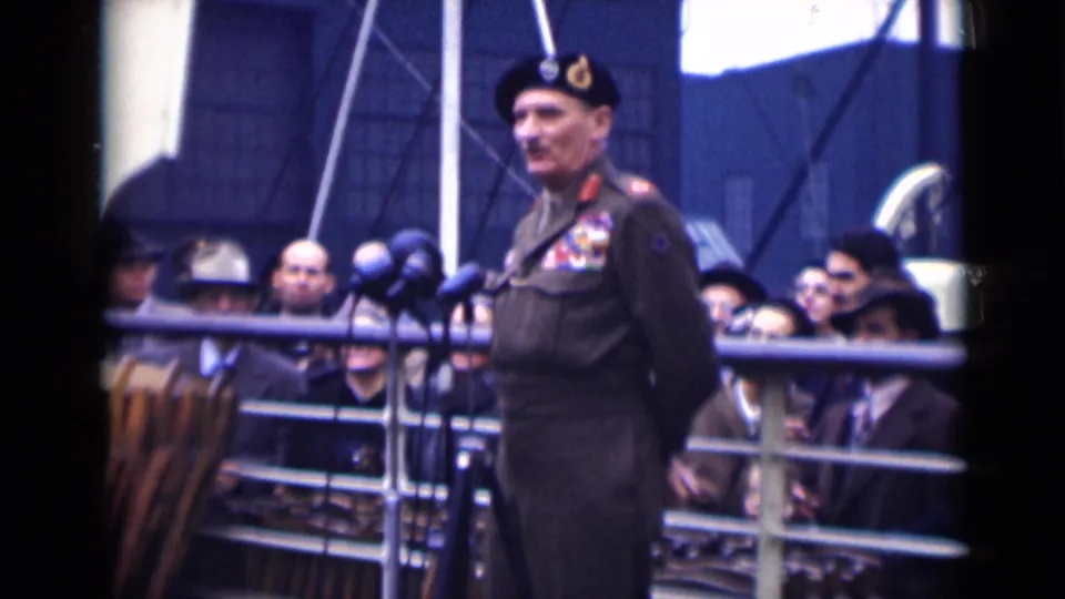 A decorated officer in uniform appears beside several microphones on a deck, with London 1946 spectators visible behind him.