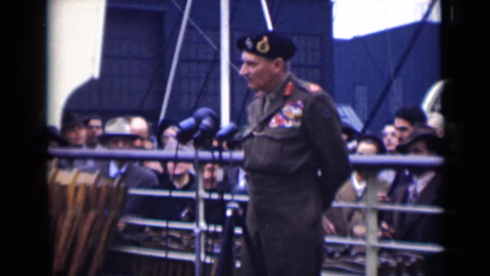 The officer turns in profile near the microphones as spectators in coats and hats watch from behind the deck railings.