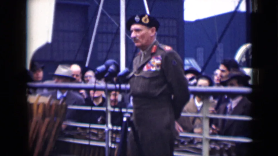 In London 1946 footage, a uniformed man with medals stands at clustered microphones while civilians watch from behind a metal railing.