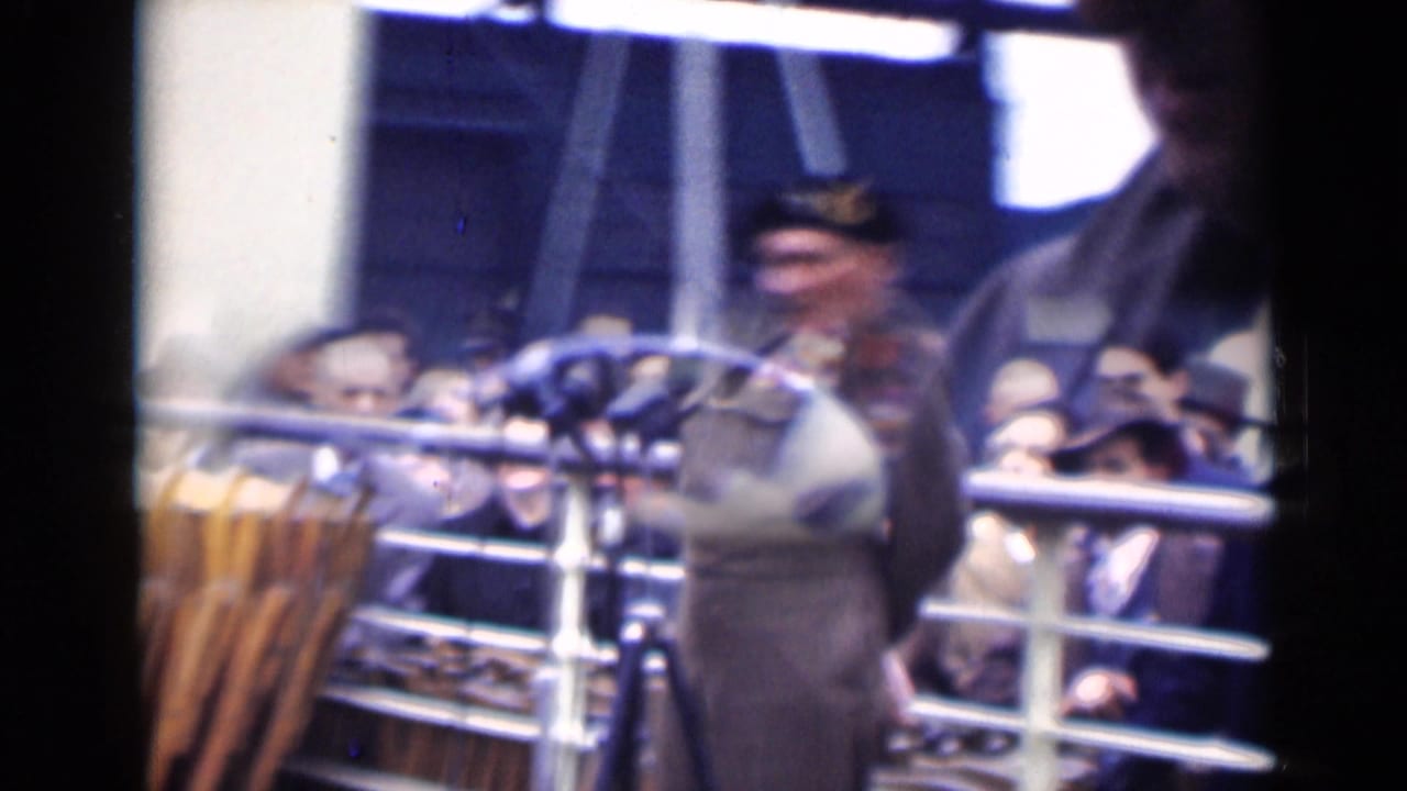 A soft 1940s London frame shows the uniformed speaker near microphones, with spectators in hats visible behind railings.