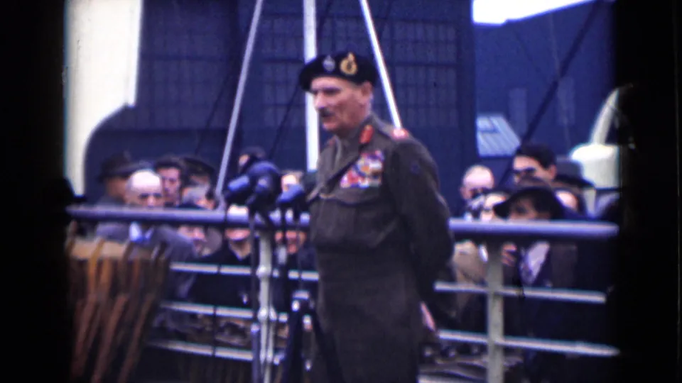 The speaker appears closer in frame, wearing a beret and decorated uniform while civilians look on behind the railing.