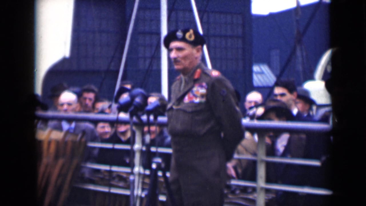 Uniformed military officer stands at microphones before a crowd behind railings.