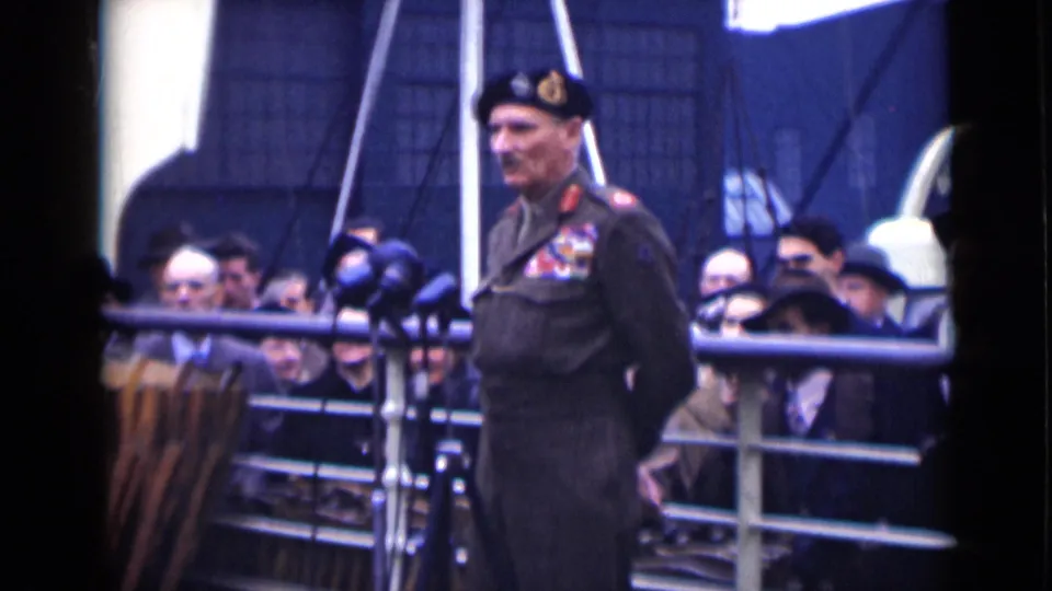 A beret-wearing military officer with medals stands near microphones as a London crowd watches from behind railings.
