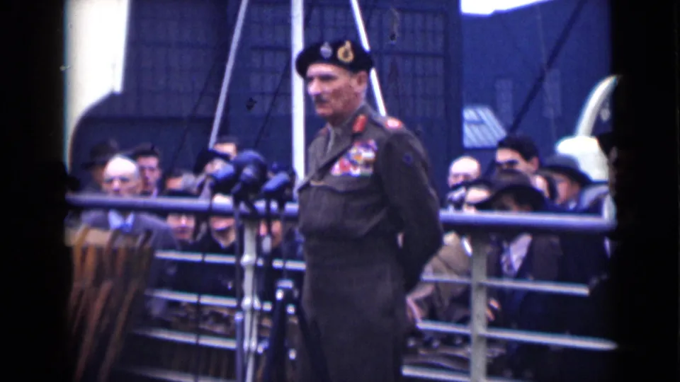 The 1946 London home-movie frame shows a decorated officer standing stoically at microphones before civilians in period coats and hats.