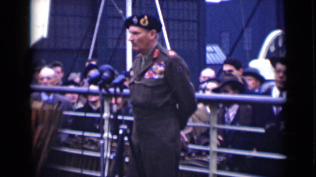 In London archival footage, a decorated officer in beret stands at microphones with spectators behind a railing.