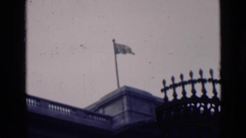 A rooftop flag and stone balustrade are framed by dark vertical edges and decorative ironwork.