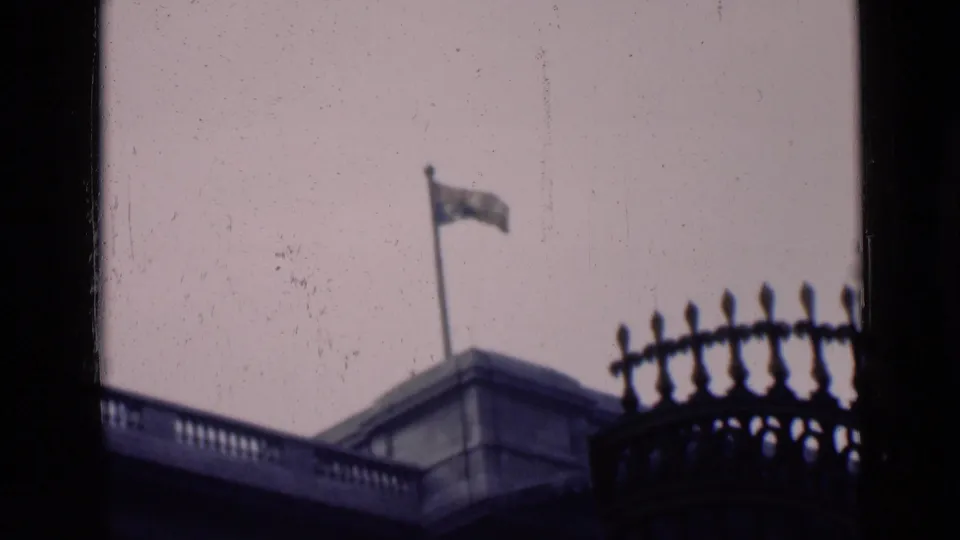 1946 London home-movie frame showing a flag atop a stone building, viewed through a window with ornate ironwork in the foreground.