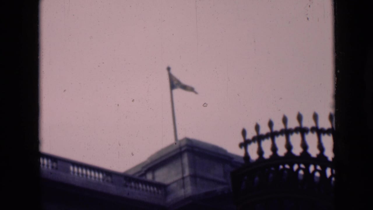 1946 London frame with a rooftop flagpole and waving flag above stone architecture, seen from a partially obstructed home-movie viewpoint.