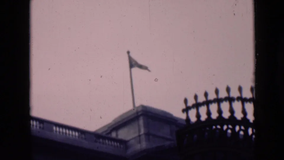 1946 London frame with a rooftop flagpole and waving flag above stone architecture, seen from a partially obstructed home-movie viewpoint.