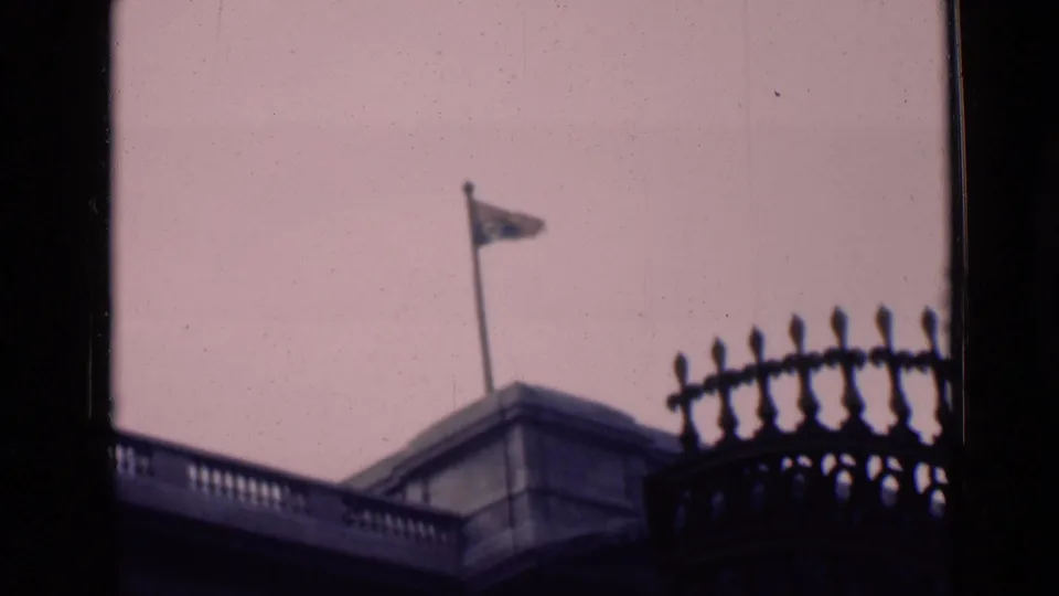 Soft 1946 London 8mm frame showing a flag over a stone roofline, with silhouetted iron railing and window edges in front.