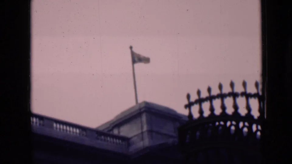 1946 London home-movie frame of a flag flying above stone architecture, with window framing and ornate ironwork partially obscuring the view.