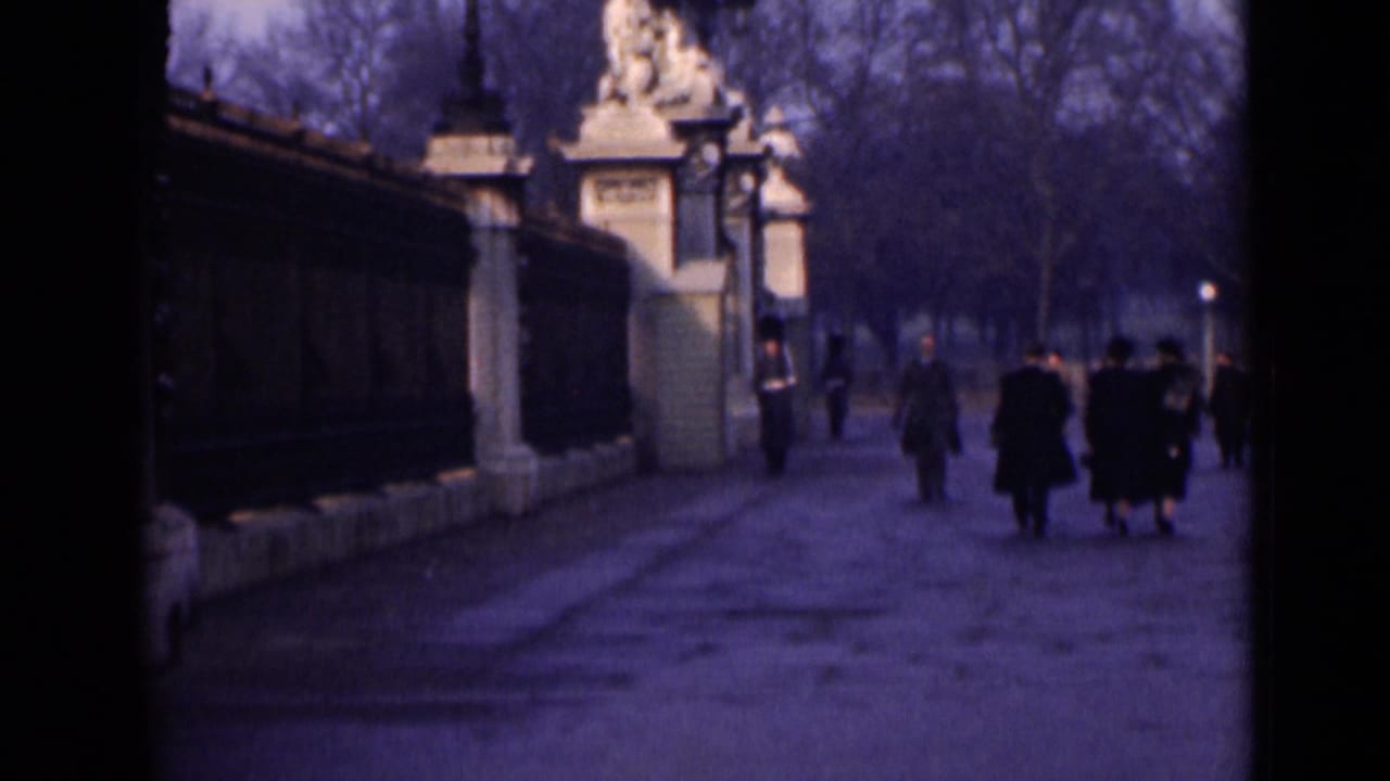 1946 London home-movie frame showing pedestrians on a broad path beside palace railings and a guard near ornate stone gates.