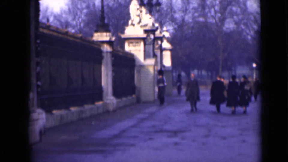 1946 London frame with a ceremonial guard beside palace gates and small groups of pedestrians walking under bare trees.
