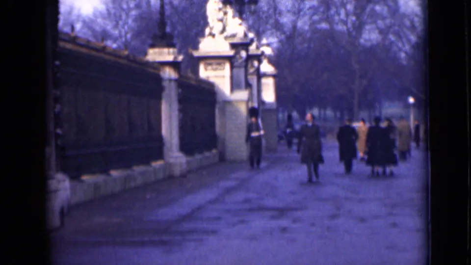 1946 London home-movie view of walkers in period coats passing palace railings, stone gateposts, and a uniformed guard.