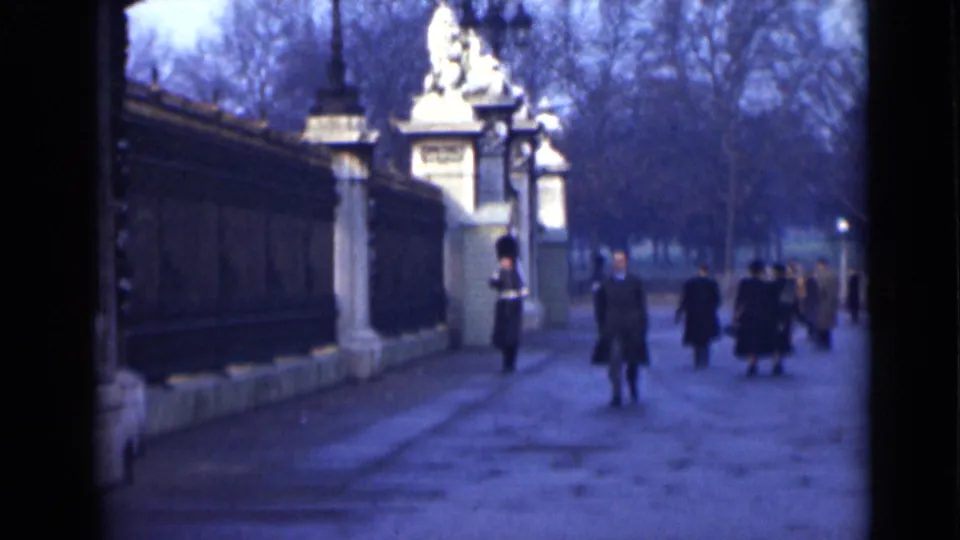 1946 London frame showing a pedestrian approaching on the path, with palace gates, railings, and a guard behind them.