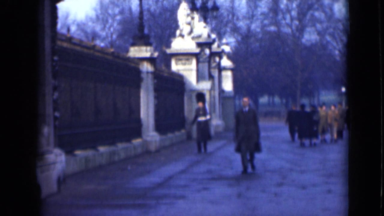 A man in a dark overcoat walks toward the camera near a guarded gate.