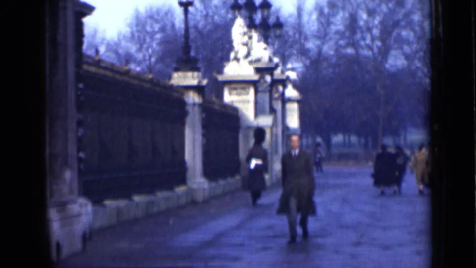 1946 London home-movie frame showing a man in an overcoat approaching, with a uniformed guard and pedestrians near an ornate entrance.