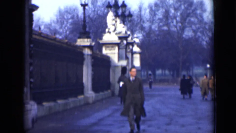 1946 London home-movie frame with a man near the foreground, pedestrians behind him, and an ornate guarded entrance in soft focus.