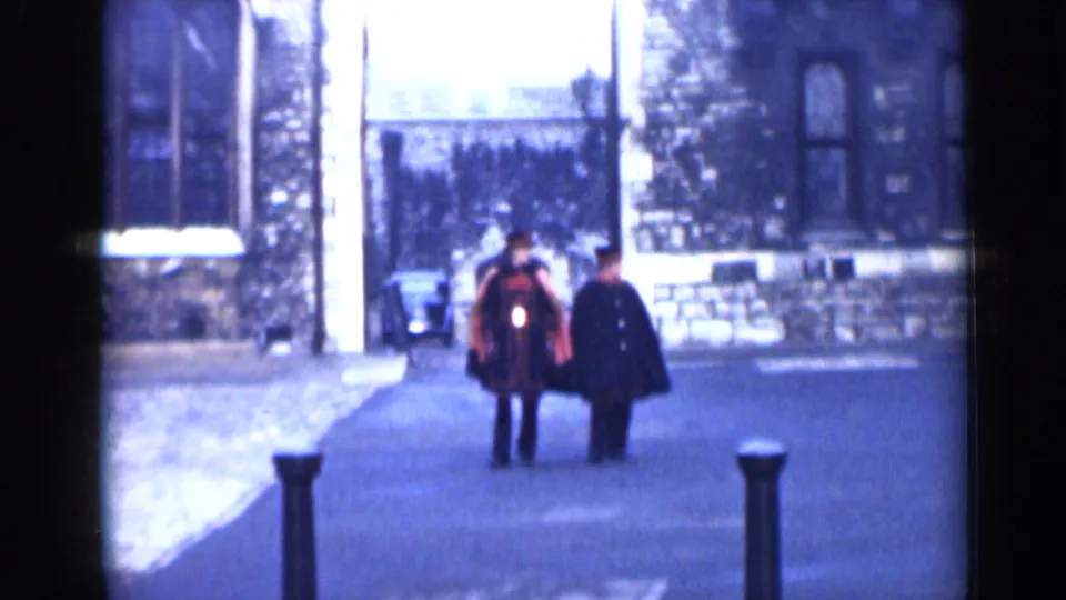 London 1946 street view with two adult men walking toward the camera, one in a red and black uniform, near stone walls and bollards.