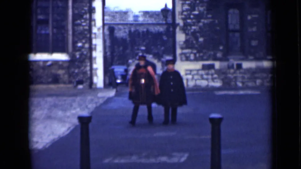 London 1946 home-movie frame of two adult men walking near stone walls and bollards, with one wearing a ceremonial-style red uniform.