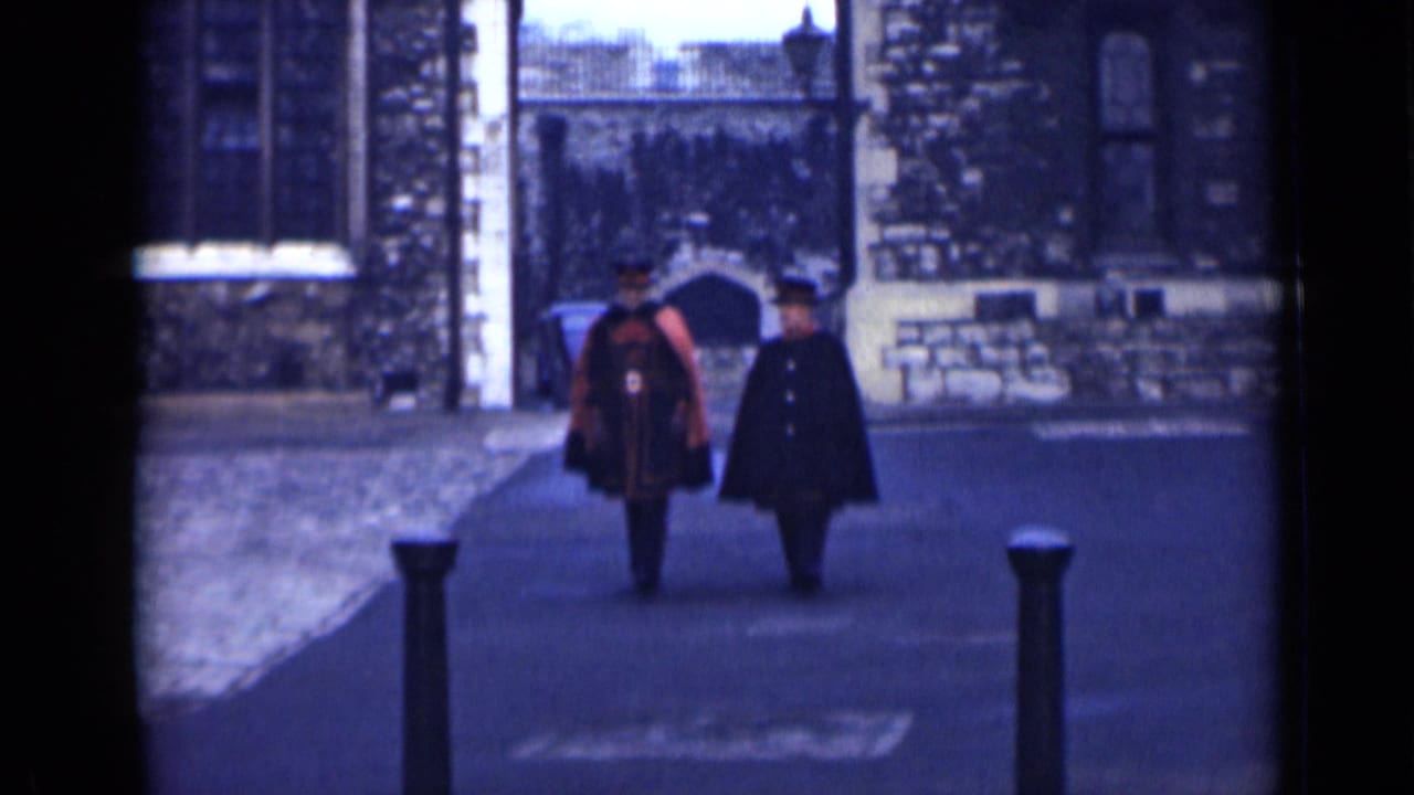 In London, England, 1946, two adult men walk side by side through a stone courtyard, one in red ceremonial dress and one in a dark coat.